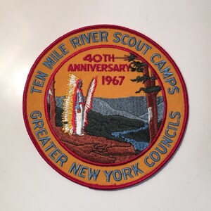 Puede incluir: Un parche bordado en amarillo y rojo con el texto "Ten Mile River Scout Camps Greater New York Councils 40th Anniversary 1967" y una representación de una figura nativa americana frente a un paisaje montañoso.