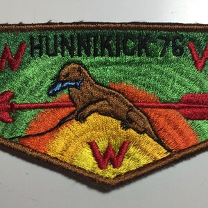 May include: A brown and green embroidered patch featuring a brown otter holding a blue fish in its mouth. The otter is standing on a yellow and orange mountain with a red arrow passing through it. The patch is inscribed with the text "W HUNNIKICK 76 W" in black lettering.