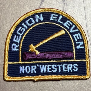 May include: A navy blue embroidered patch with gold stitching. The patch features a hammer and log with the text "REGION ELEVEN NOR'WESTERS" in white.