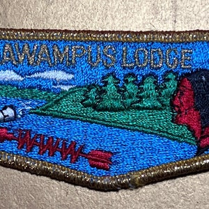 Puede incluir: Un parche bordado azul y verde con el texto "Tonkawaumpus Lodge 1A". El parche presenta una canoa con dos figuras en ella, una pequeña isla con árboles y una figura nativa americana con un tocado de plumas.