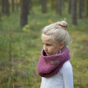 May include: A young girl with blonde hair wearing a pink knitted neck warmer with a brown label that says "Walden". The girl is standing in a forest.