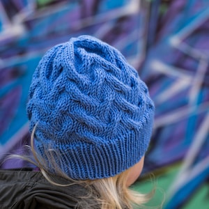 May include: A blue knitted hat with a cable knit pattern. The hat is worn by a person with blonde hair.