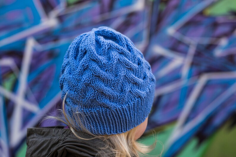 May include: A blue knitted hat with a cable knit pattern. The hat is worn by a person with blonde hair.