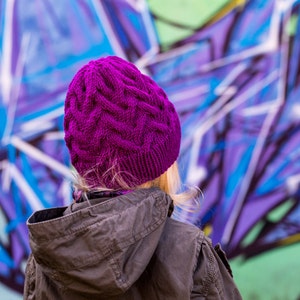 May include: A person wearing a purple knitted hat and a brown jacket, standing in front of a graffiti wall with purple, blue, and white paint.