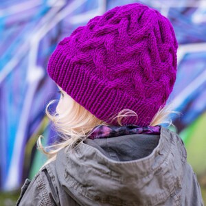 May include: A young person wearing a purple knitted hat with a cable knit pattern. The hat is worn over their blonde hair. The person is wearing a gray hooded jacket.