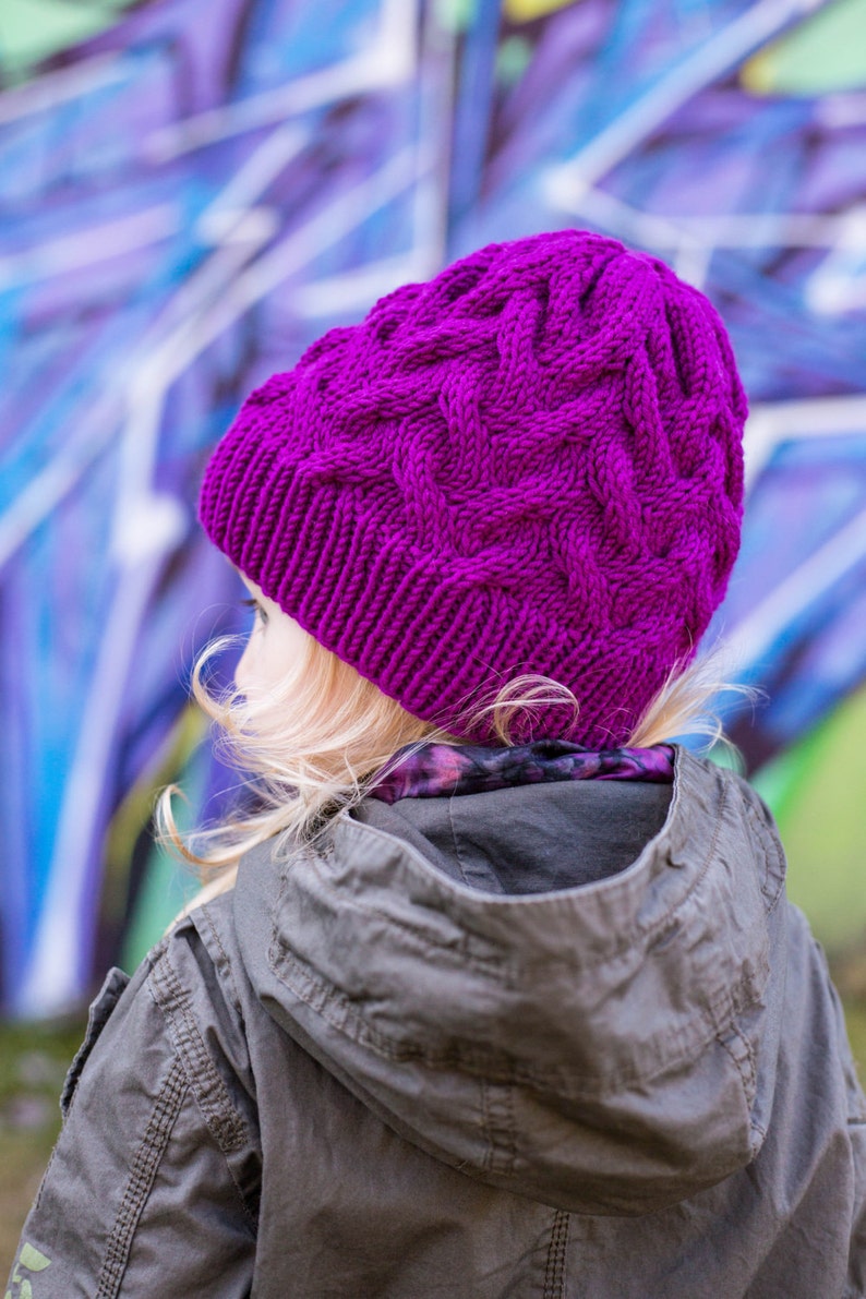 May include: A young person wearing a purple knitted hat with a cable knit pattern. The hat is worn over their blonde hair. The person is wearing a gray hooded jacket.