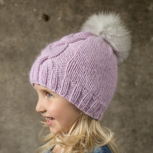 May include: A young girl wearing a purple knitted hat with a white fur pom pom. The hat has a cable knit pattern. The girl has long blonde hair and is wearing a denim jacket.