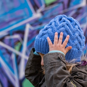 May include: A young person wearing a blue knitted hat with a cable knit pattern. The hat is pulled down over their forehead and they are holding it with one hand. They are wearing a dark gray jacket.
