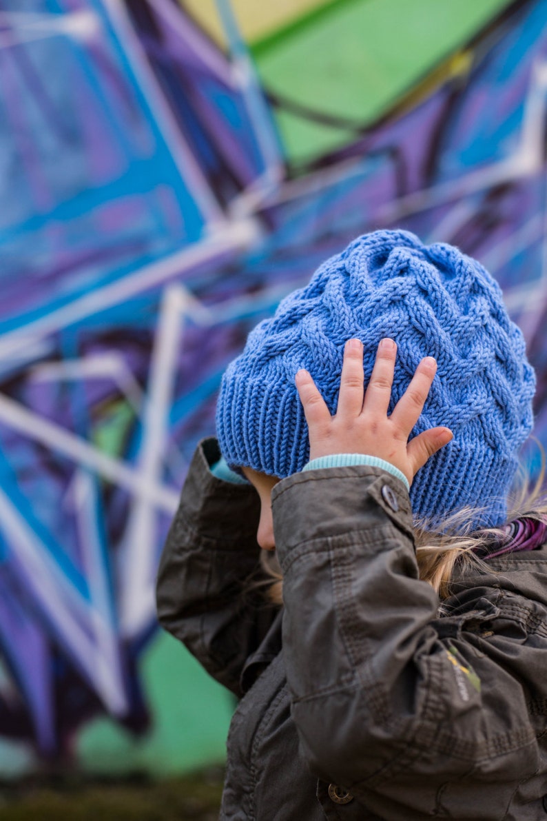 May include: A young person wearing a blue knitted hat with a cable knit pattern. The hat is pulled down over their forehead and they are holding it with one hand. They are wearing a dark gray jacket.