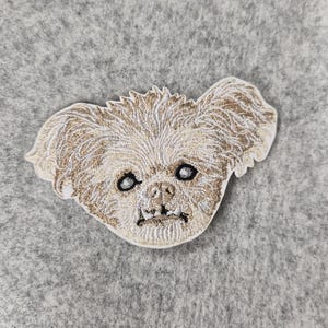 May include: An embroidered patch featuring the head of a small, shaggy dog with light tan and cream-colored thread. The dog has large, dark eyes, a small nose, and visible lower teeth, with a textured, stitched appearance on a white border.