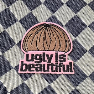 May include: An embroidered patch featuring a brown bowl-cut hairstyle graphic above the black text "ugly is beautiful" on a light pink background. The patch has a pink border and is displayed against a dark blue and white checkered fabric.