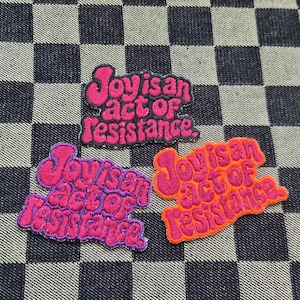 May include: Three embroidered patches with the phrase "Joy is an act of Resistance." The patches are in pink, purple, and orange, with a black and white checkered background.