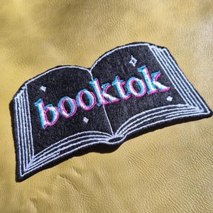 May include: Embroidered patch featuring an open book design with the word "booktok" in pink and blue lettering. The patch is black with white stitching and is set against a yellow background.