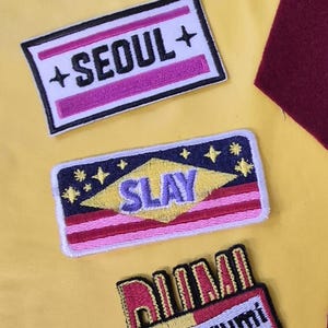 May include: A collection of embroidered patches on a yellow background. The patches feature text and designs, including "HUNTRIX World Tour" in blue, "+SEOUL+" in pink and white, and "SLAY" with stars, plus a red and yellow patch.