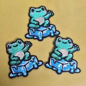 May include: Three embroidered patches featuring a cartoon frog waving from a blue ice block. The frogs are teal with tan bellies and black outlines. The ice blocks are light blue with darker blue shading and black outlines. The background is yellow.