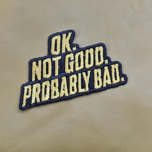 May include: Embroidered patch with the phrase "OK. NOT GOOD. PROBABLY BAD." in gold thread on a navy blue background. The patch is rectangular with a slight curve and is set against a tan background.