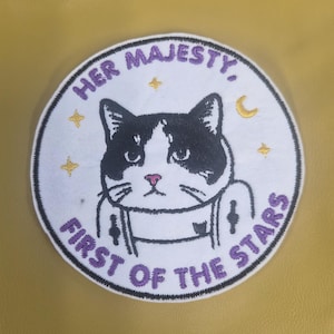 May include: Embroidered patch featuring a black and white cat with the text "HER MAJESTY, FIRST OF THE STARS" in a circular design. The patch includes star and crescent moon accents, set against a yellow background.