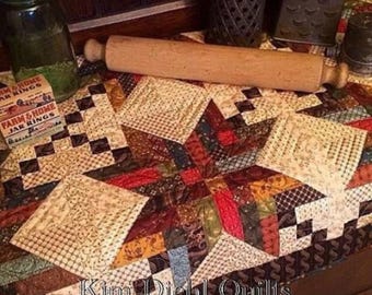 May include: A patchwork quilt featuring a central star pattern in shades of red, brown, and tan with intricate geometric fabric squares. The quilt displays the text Kim Dichl Quilts and is positioned near a wooden rolling pin and a jar ring box.