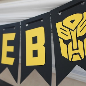May include: Black banner with yellow letters spelling "CALEB" and a yellow Transformers logo. The banner is strung across a white surface. The letters and logo are bold and easily readable.