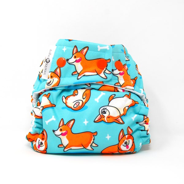 May include: A blue cloth diaper with a pattern of orange and white corgis on a blue background. The diaper has white stars and bone-shaped designs. The text "A Little Squirrel" is printed on the diaper.