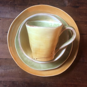May include: A set of three ceramic plates in shades of green and orange. The plates are nested together, with a yellow mug in the center.