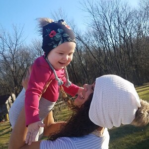 May include: A woman wearing a white knit beanie with a fluffy pom-pom holds a young child wearing a pink shirt and a floral print beanie. The child is smiling and looking up at the woman.