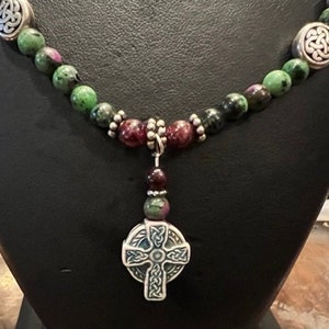 May include: A green and purple gemstone necklace with a silver Celtic cross pendant. The necklace is made with round beads and has silver accents.