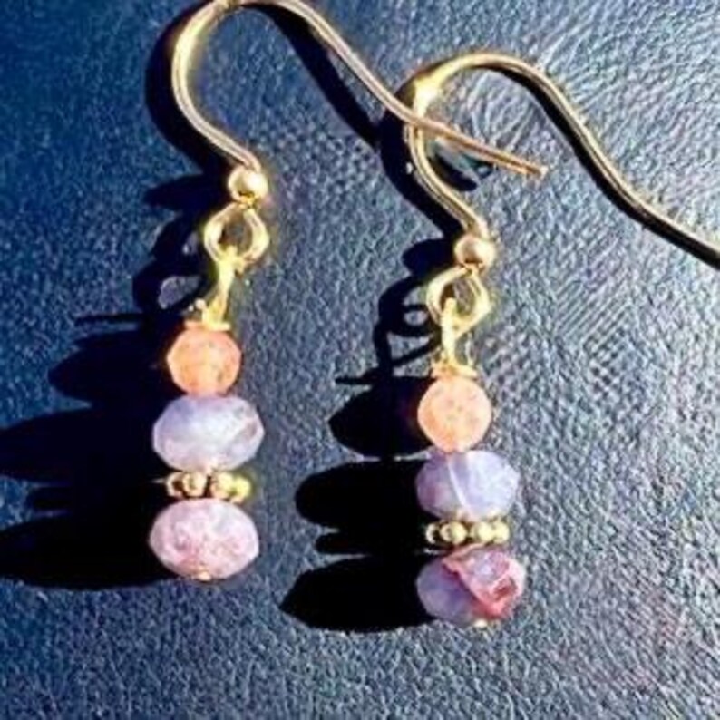 Puede incluir: Un par de pendientes dorados con dise&ntilde;o de gancho. Cada pendiente presenta una pila de tres cuentas de piedras preciosas: una cuenta color melocot&oacute;n, una cuenta color lavanda y una cuenta rosa rojiza. Los pendientes se muestran sobre un fondo azul oscuro.