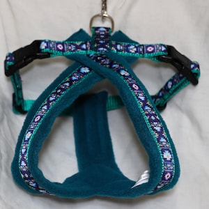 May include: Teal dog harness with a patterned ribbon trim in purple, blue, and pink. The harness features black plastic buckles and a metal ring for leash attachment. The harness is made of a soft, plush material.