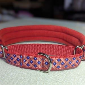 以下が含まれることがあります： パッド入りの首と模様入りのバンドが付いた赤い犬の首輪。バンドは、赤い背景に青と白の正方形を繰り返すデザインが特徴です。首輪には、シルバーの金属製のバックルとDリングが付いています。