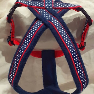 May include: A blue dog harness with white polka dots and red trim. The harness has a soft, plush texture and features adjustable black buckles and a metal ring for leash attachment. The design is a classic combination of navy, red, and white.