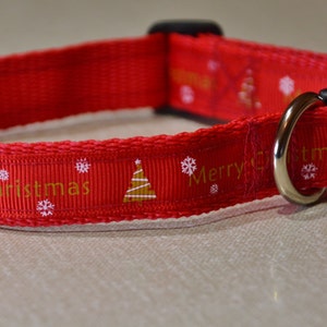 May include: Red dog collar with a white background and gold lettering that says "Merry Christmas". The collar has a black buckle and a silver D-ring.
