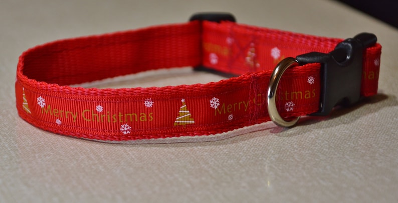 May include: Red dog collar with a white background and gold lettering that says "Merry Christmas". The collar has a black buckle and a silver D-ring.