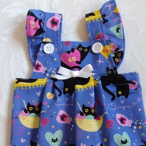 Może przedstawiać: Niebieskie szorty na szelkach z powtarzającym się wzorem czarnych kotów, serc i kłębków wełny. Paski mają marszczone krawędzie i białe guziki. Biała kokarda jest wyśrodkowana z przodu. Szorty na szelkach są dla lalki.