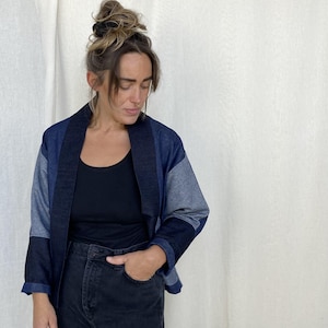 May include: A blue denim jacket with a black collar and a slightly cropped hem. The jacket is made of different shades of blue denim and has a relaxed fit.