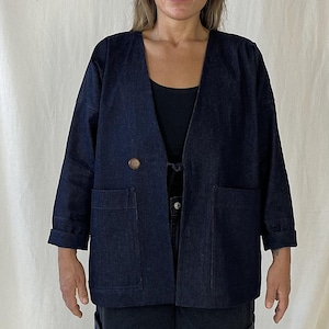 Può includere: Una giacca di jeans blu scuro con un collo largo aperto e due grandi tasche. La giacca ha una chiusura con un solo bottone e una cravatta in vita.