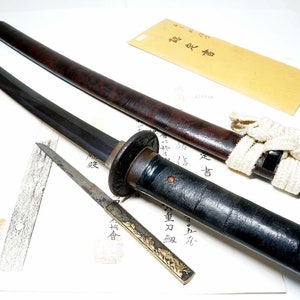 May include: A traditional Japanese sword, or katana, with a black scabbard and a brown wooden handle. The sword is displayed with a smaller knife, a white cord, and a paper document with Japanese writing.