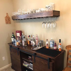 May include: A dark wood bar cabinet with sliding doors and a shelf above. The shelf has a wine glass rack and is filled with glasses. The cabinet is stocked with liquor bottles and a bottle of soda. There is a black bar mat on the counter.