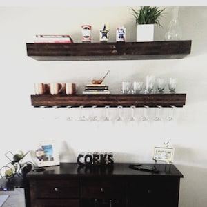 May include: A dark wood bar with two shelves, holding glasses, mugs, and decorative items. Below, a cabinet with the word "CORKS" and a wine rack. The bar is against a white wall, creating a clean, modern look.