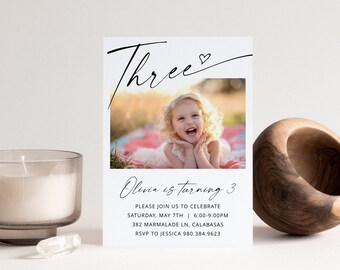 Op de afbeelding: Een witte verjaardagsuitnodiging met zwarte kalligrafie die "Three" zegt met een klein hartje. De uitnodiging heeft een foto van een jong meisje met blond haar dat lacht. De tekst op de uitnodiging luidt "Olivia wordt 3" en bevat de datum, tijd, adres en RSVP-informatie.