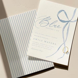 Könnte beinhalten: Eine cremefarbene Einladungskarte mit einer blauen Aquarell-Schleifenillustration, einer goldenen Ringgrafik und dem Text Something Blue Before I Do, Emily Mayers sowie Veranstaltungsdetails, dazu ein passender Umschlag mit blauen vertikalen Streifen.