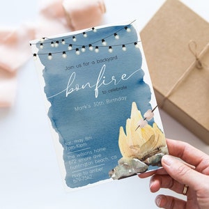 May include: A blue and white invitation for a backyard bonfire to celebrate a 30th birthday. The invitation features a watercolor background with string lights and a campfire with marshmallows. The text reads "Join us for a backyard bonfire to celebrate Mark's 30th Birthday. Sat, May 5th 7pm-10pm The Wilsons home 672 Smore Ave Huntington Beach, CA RSVP to Amber 679-7540."