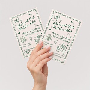 May include: Two cream-colored birthday invitations with green text and illustrations are held by a hand. The invitations read "She's not that Matcha older" and include details for Sophia's 18th birthday celebration.