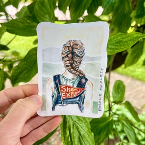 May include: A watercolor illustration of a person with long braided hair wearing a backpack with a red and white flag that says "She Explores". The background is a watercolor wash of blue, green, and gray.