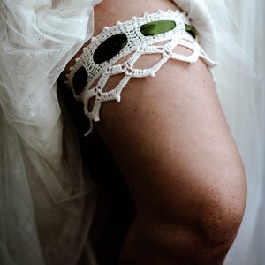 May include: A white crochet garter featuring an intricate openwork pattern and a dark green ribbon woven through the upper section. The garter is positioned on a leg, partially covered by layers of sheer white fabric with a delicate lace texture.