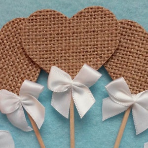 Könnte beinhalten: Sechs braune Cupcake-Topper aus Jute in Herzform mit weißen Satinbändern. Die Topper sind an Holzspießen befestigt.