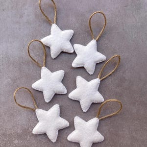May include: Six white felt star ornaments, each featuring a five-pointed shape and a brown jute twine loop attached to the top for hanging. The stars have a soft, textured surface and are arranged in a scattered pattern.