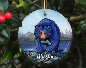 May include: A round ceramic ornament featuring a blue bear with a salmon in its mouth. The bear is standing in a stream with mountains in the background. The text "Alaska" is written below the bear.