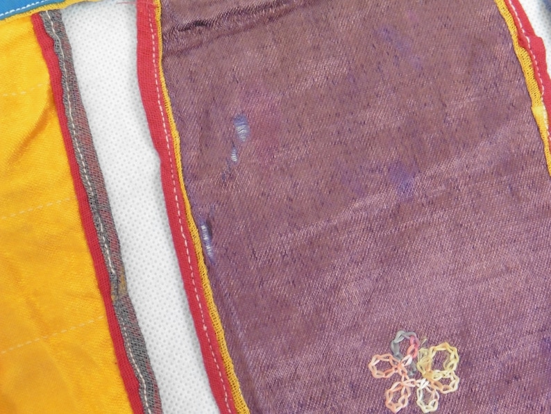 Puede incluir: Primer plano de una tela morada con un dise&ntilde;o de flor bordada en amarillo y rosa. La tela est&aacute; bordeada con ribetes rojos, amarillos y grises.