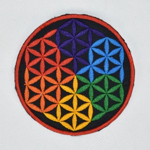 May include: A round, black embroidered patch featuring a colourful flower of life design. The design is made up of overlapping circles and triangles in shades of red, orange, yellow, green, blue, and purple.